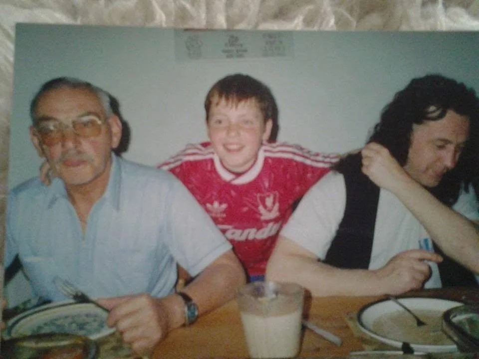 A young Luke in a red football shirt sitting between his father and grandfather at a table