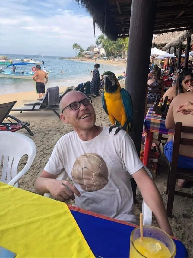 Luke smiling on a Puerto Vallarta beach with a blue-and-gold parrot perched beside him